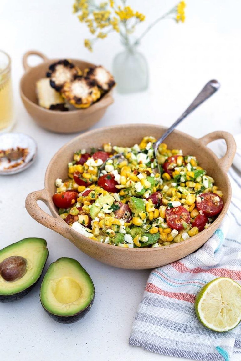 corn tomato avocado salad in neutral colored bowl with avocado and lime next to it