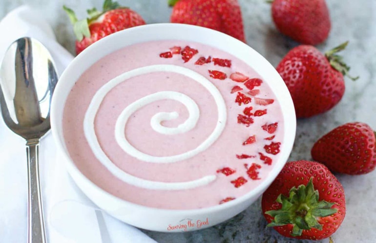 Strawberry soup with white garnish in white bowl with spoon and whole strawberries next to it