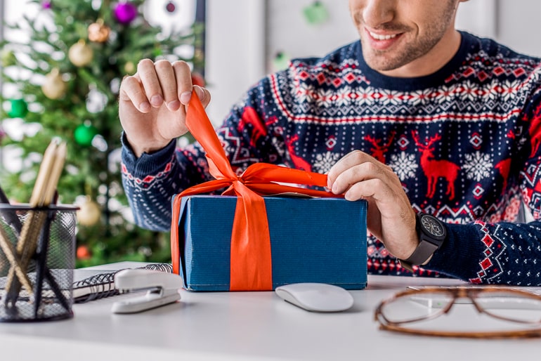 Man in christmas sweater unwrapping gift with blue paper and orange ribbon on desk