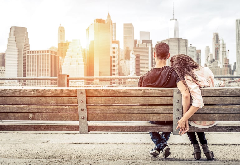 Couple sitting on park bench holding hands with city buildings in background