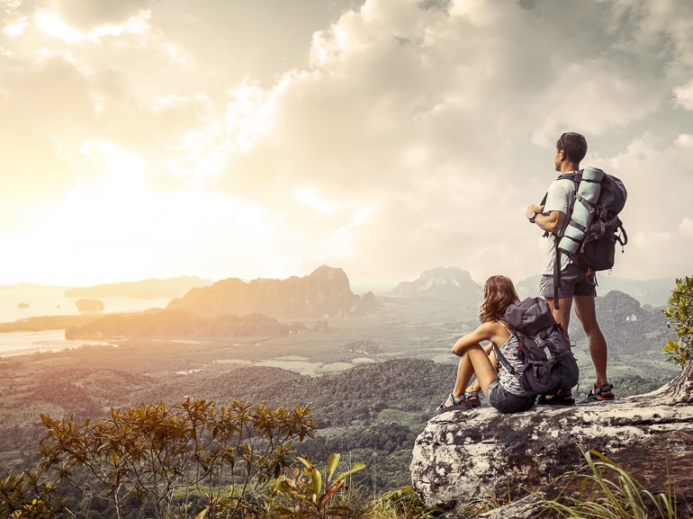 couple with backpacks standing on edge of cliff with sun and views
