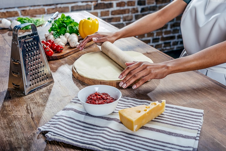 Person rolling out dough on wooden board with vegetables and cheese next to it