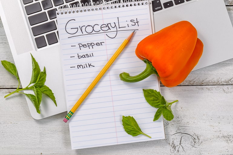 Orange pepper and pencil laying on top of grocery list and laptop