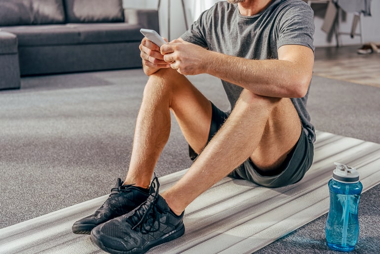 Man sitting on workout mat in living room with phone and water bottle