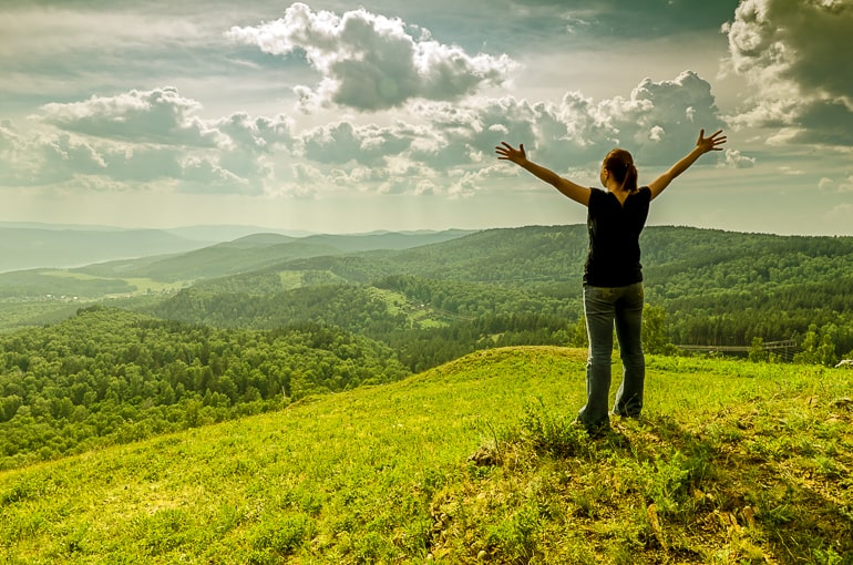 Woman standing on hill with raised arms and green forest in the background
