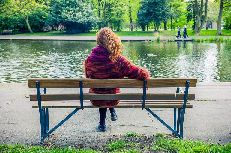 Photo of woman sitting on brown bench with lake and pathway in the background