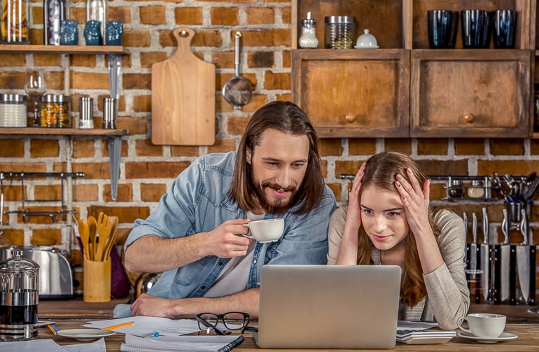 Man and woman in kitchen looking at laptop screen with coffee next to them
