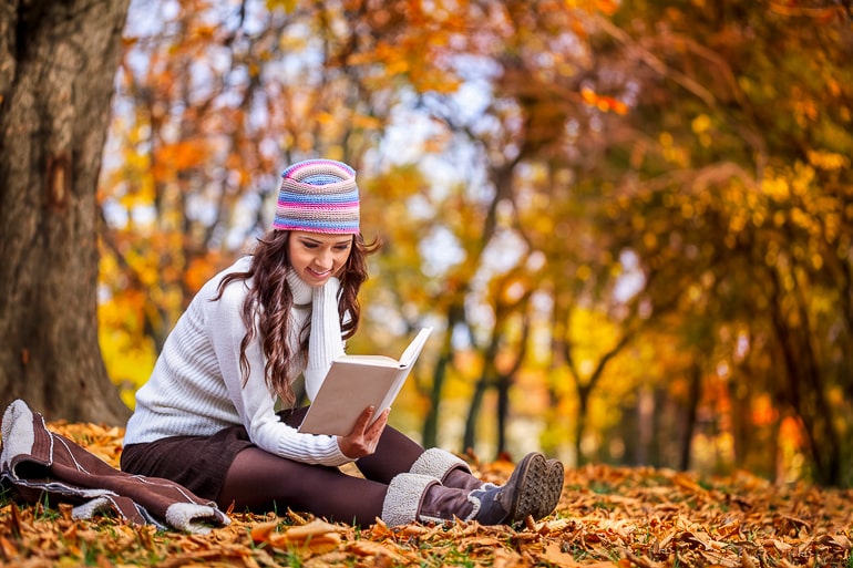 Woman with hat sitting on ground covered in fall leaves and reading a book