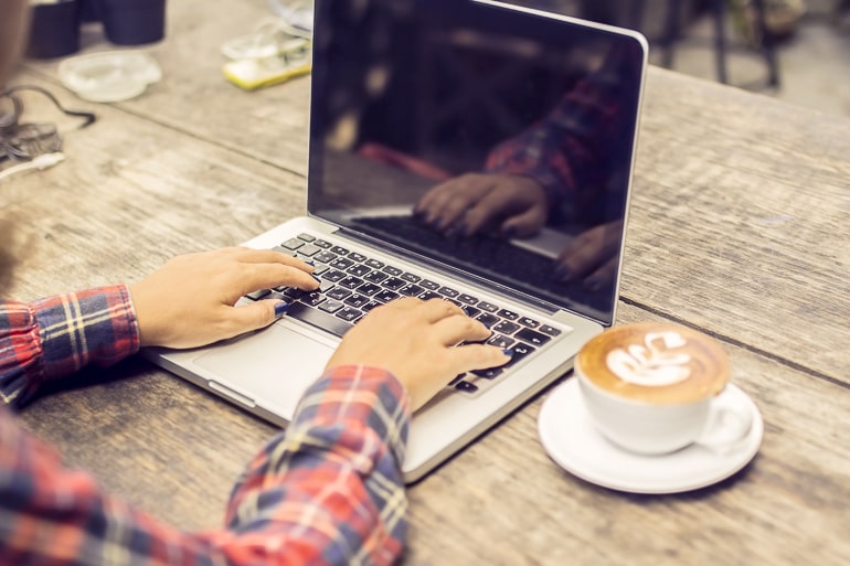 Person typing on computer on wood desk with cappuccino next to it