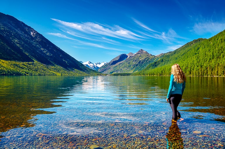 Woman with blonde hair standing in clear water with mountains and forest in background