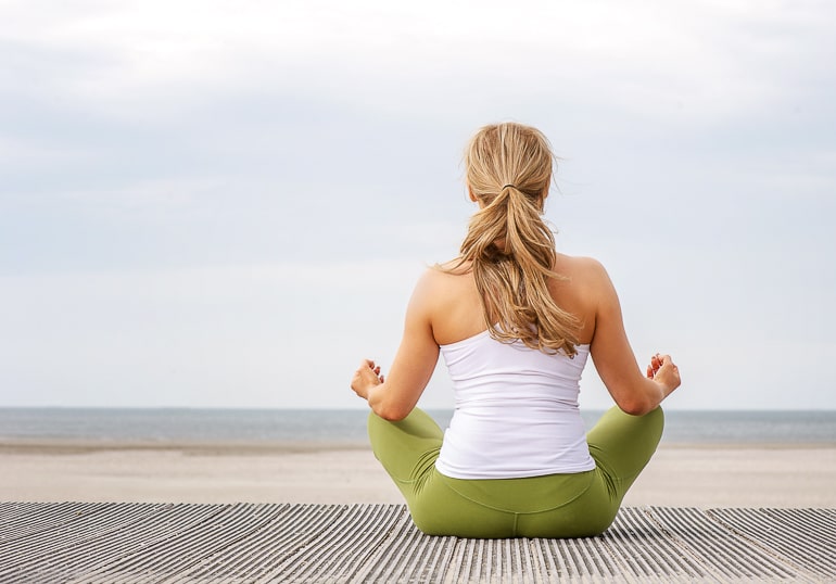 woman with green pants sitting in yoga pose looking at ocean living in the present