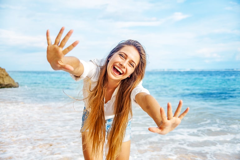 woman with long hair waving and smiling at camera with beach behind