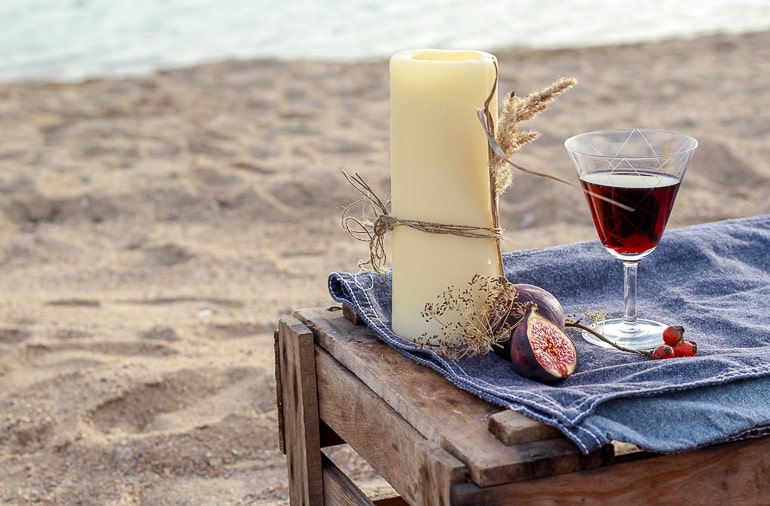 Candle with fruit and glass of red liquid on crate with sand and water in background