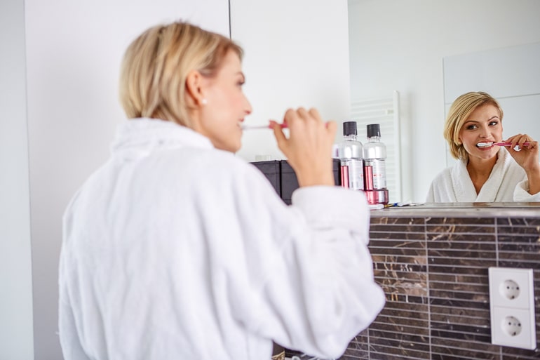 Woman in white robe brushing teeth and looking in mirror