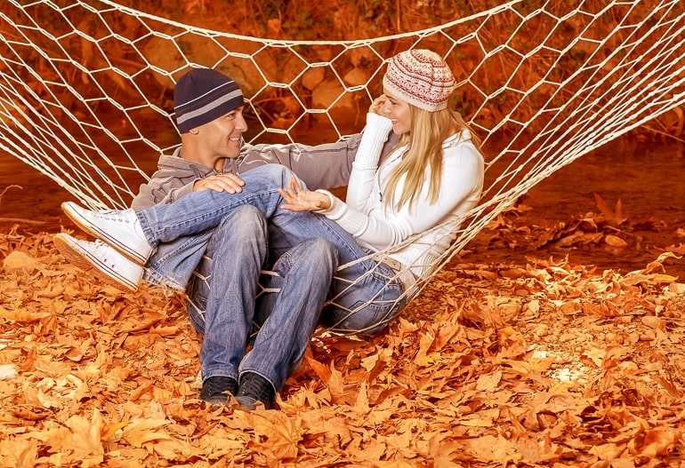 Man and woman sitting in white hammock looking at each other with fall leaves on ground