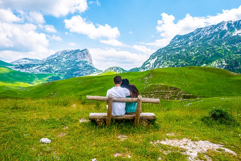 Couple sitting on wood bench with green grass and tall mountains in background self care