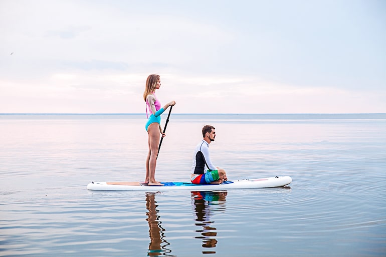 Man sitting on Stand Up Paddleboard with woman standing behind him on lake