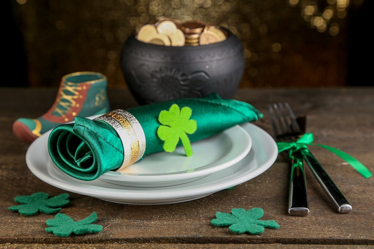 green rolled napkin in white plate with pot of gold behind on wooden table