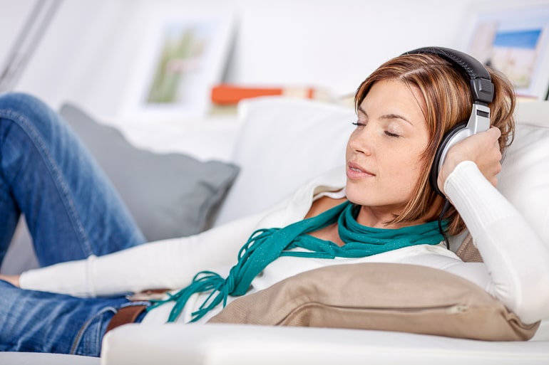 Woman with eyes closed sitting on white couch and listening to music over headphones