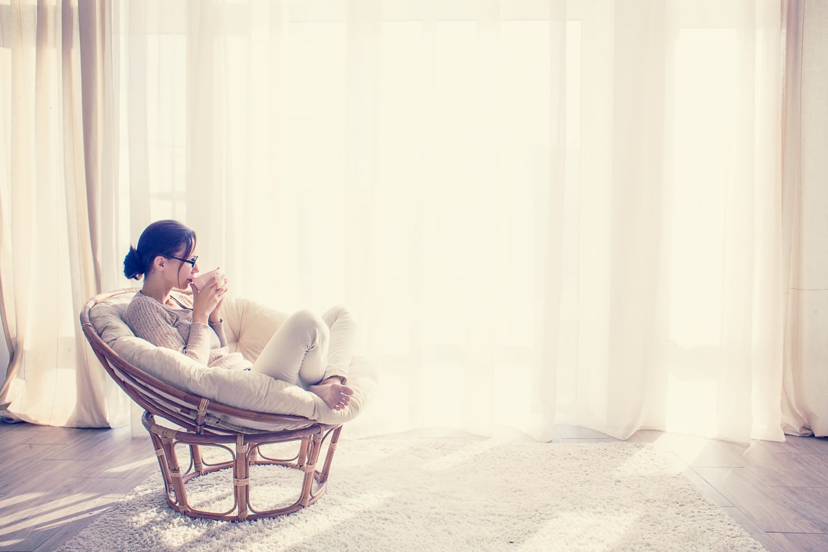 Woman sitting in comfortable chair and holding a mug in front of white curtains