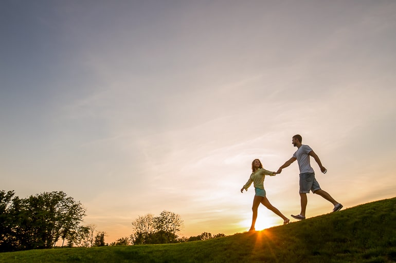 Man and woman walking on grass and holding hands with setting sun in background
