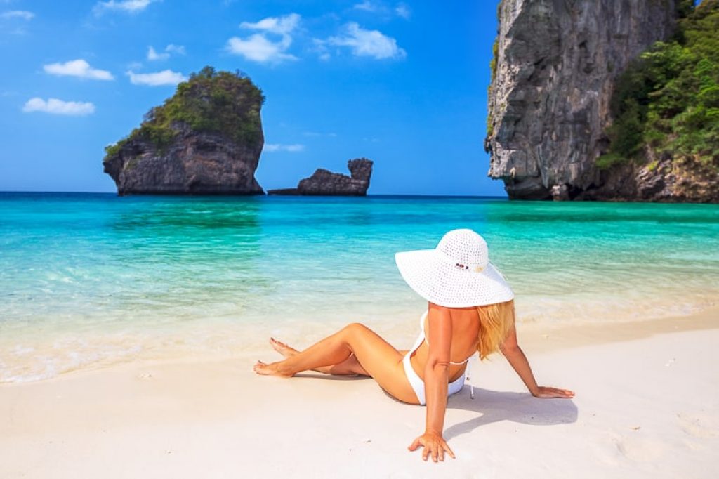 Woman in white bikini with white hat sitting on sandy beach with clear water and cliffs in background