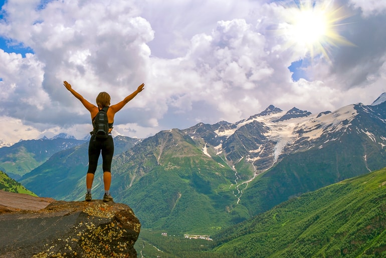 Woman Hiker standing on rock raising her arms with sun and mountains in background