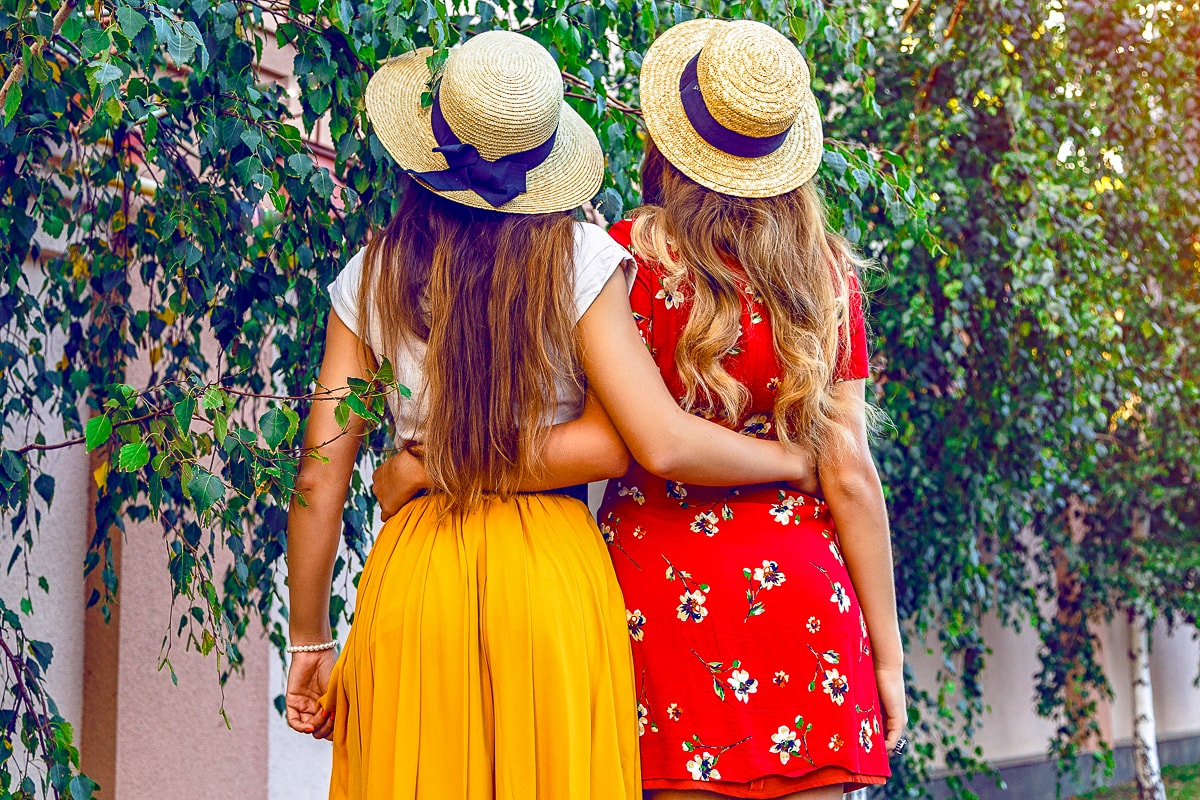 Two women with long hair and hats wrapping their arms around each other with greenery in background best friend captions