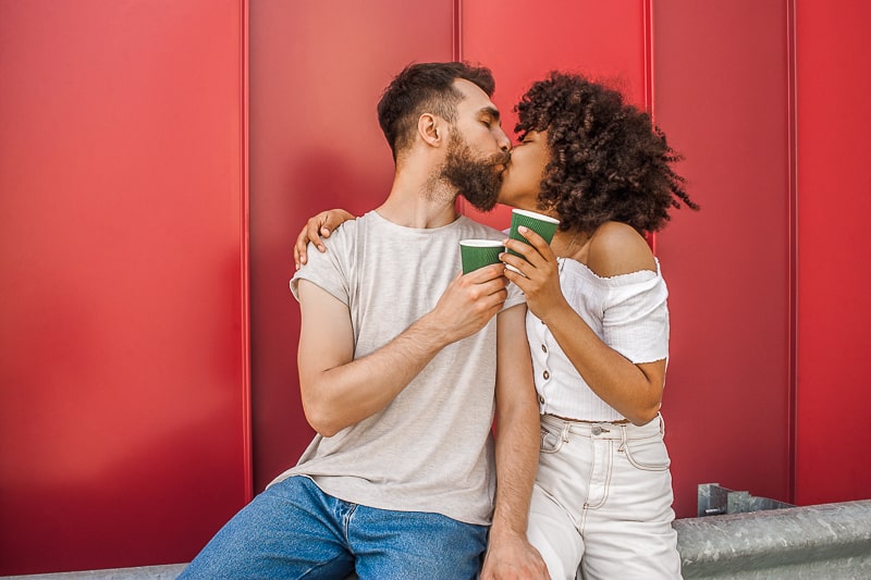 man and woman kissing holding coffees with red backdrop