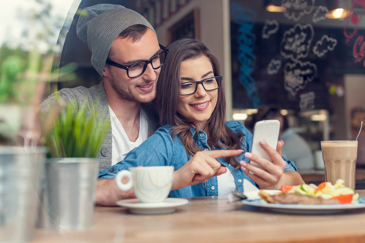 Couple sitting in a cafe with food in front of them looking at a smartphone apps for couples