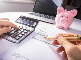 Hands holding a pen and typing on a calculator with pink piggy bank and computer in background benefits of budgeting