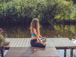 Woman sitting cross legged on wooden deck with water and greenery in background