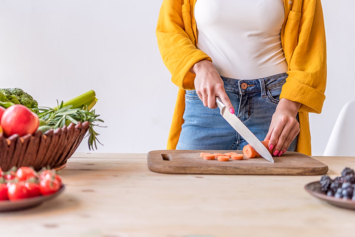 Woman cutting carrot on brown cutting board with bowls of fruit and vegetables next to it how to eat healthy on a budget
