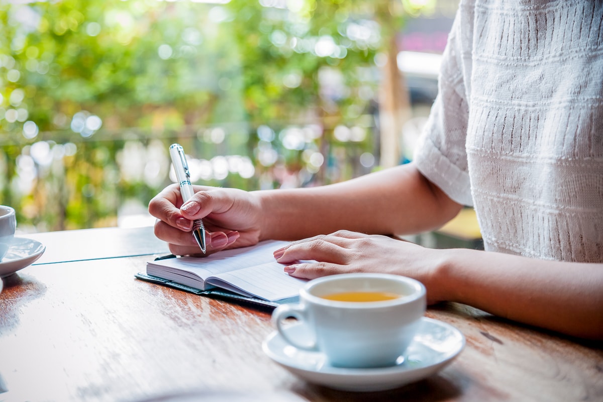 Woman writing in journal with mug of tea on table next to her how to keep a journal